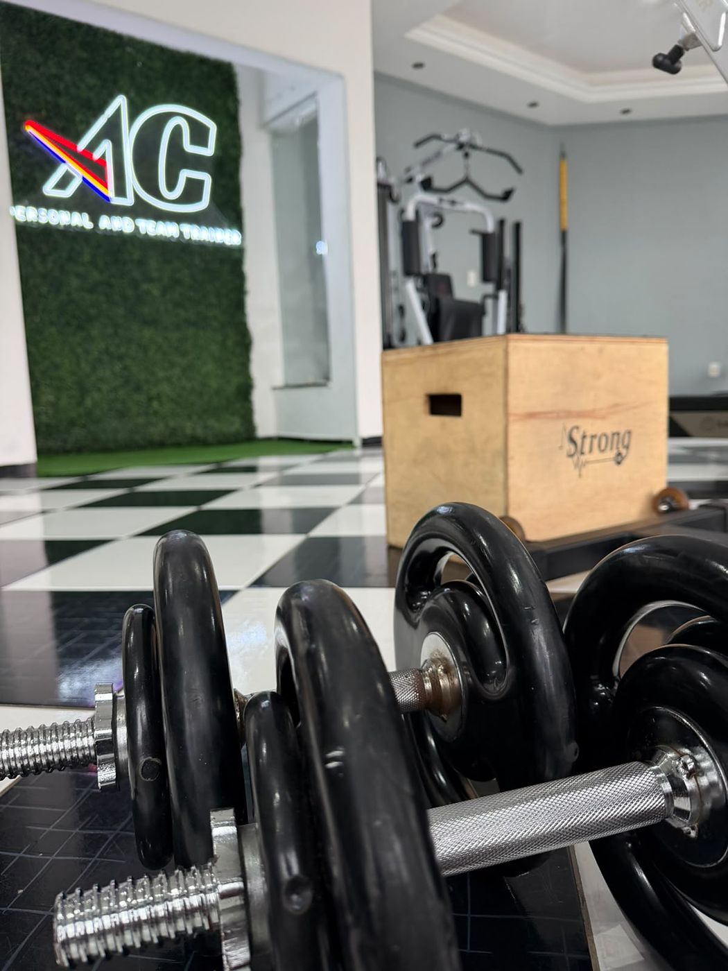 Interior do estúdio AC Personal and Team Trainer, com halteres em primeiro plano e o letreiro AC na parede verde ao fundo